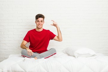 Young teenager student man on the bed holding something little with forefingers, smiling and confident.