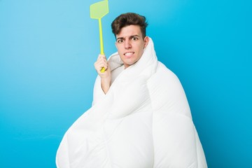 Naklejka premium Teenager caucasian tired man covering himself with a blanket and holding a flyswatter