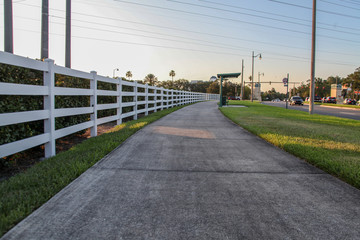 A long, curved white wooden fence alongside a grassy walk in the background of the beautiful landscape of plants and trees in the late afternoon in Celebration, Orlando, Florida, USA.