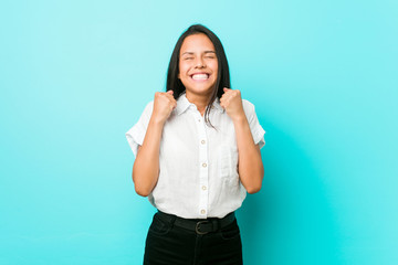 Young hispanic cool woman against a blue wall raising fist, feeling happy and successful. Victory concept.