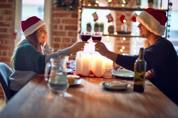Young beautiful couple smiling happy and confident. Eating food and toasting with cup of wine  wearing santa claus hat celebrating christmas at home