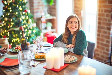 Young beautiful woman sitting eating food around christmas tree at home looking away to side with smile on face, natural expression. Laughing confident.