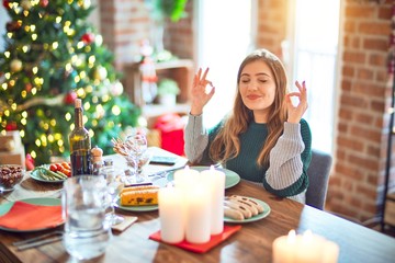 Young beautiful woman sitting eating food around christmas tree at home relax and smiling with eyes...