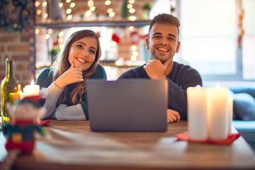 Young beautiful couple sitting using laptop around christmas decoration at home looking confident at the camera smiling with crossed arms and hand raised on chin. Thinking positive.