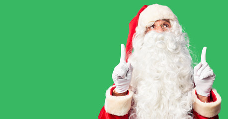 Middle age handsome man wearing Santa Claus costume and beard standing amazed and surprised looking up and pointing with fingers and raised arms.