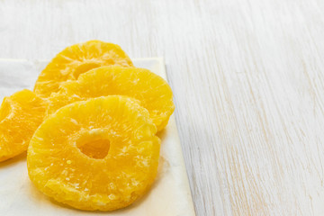 Slices of dried pineapple lie on a food paper on a white wooden background, side view from above