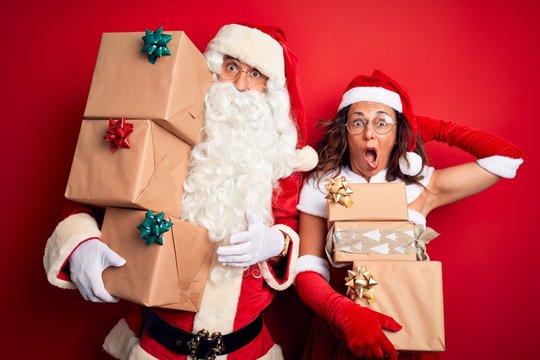 Middle age couple wearing Santa costume holding tower of gifts over isolated red background Crazy and scared with hands on head, afraid and surprised of shock with open mouth
