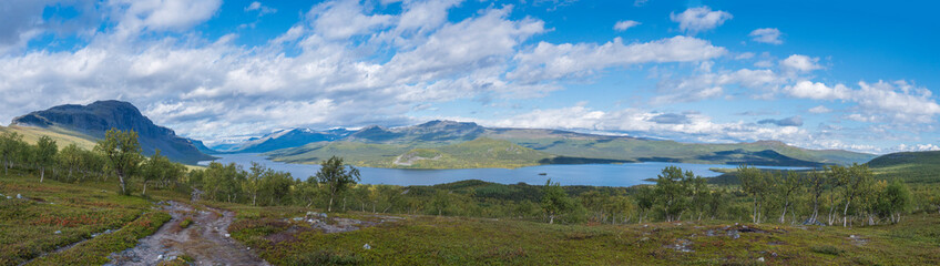 Obraz premium Panoramic landscape with beautiful river Lulealven, snow capped mountain, birch tree and footpath of Kungsleden hiking trail near Saltoluokta, north of Sweden, Lapland wild nature. Summer blue sky