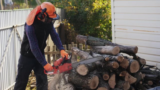 A lumberjack guy in a protective helmet cuts logs with a chainsaw in the backyard.