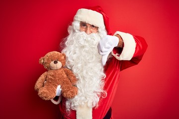 Middle age man wearing Santa Claus costume holding teddy bear over isolated red background with angry face, negative sign showing dislike with thumbs down, rejection concept
