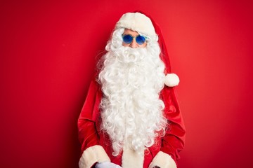 Middle age man wearing Santa Claus costume and sunglasses over isolated red background with serious expression on face. Simple and natural looking at the camera.