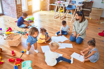 Beautiful teacher and group of toddlers sitting on the floor drawing using paper and pencil around lots of toys at kindergarten