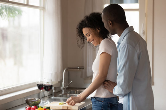 Happy man cuddling cheerful black woman preparing food.