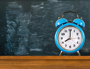An alarm clock stands on a table against the background of a blackboard with traces of chalk