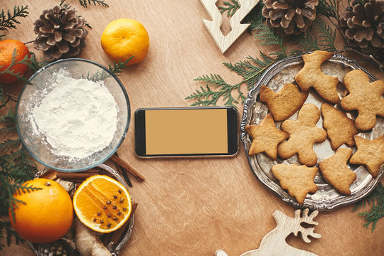 Phone with empty screen and festive gingerbread cookies with anise, cinnamon, pine cones, cedar branches on rustic table, flat lay. Space for text.Merry Christmas. Holiday card mockup
