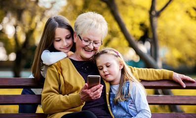 Happy family. Grandmother sitting in the park with granddaughters and having fun with smartphone....