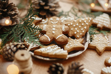 Christmas gingerbread cookies on vintage plate and anise, cinnamon, pine cones, cedar branches  with golden lights on rustic table. Baked traditional gingerbread man, tree, star cookies - obrazy, fototapety, plakaty