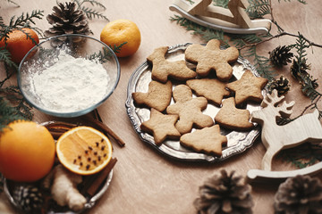Fototapeta na wymiar Christmas gingerbread cookies on vintage plate and anise, ginger, cinnamon, pine cones, fir branches, flour, oranges on rustic table. Baking traditional gingerbread cookies