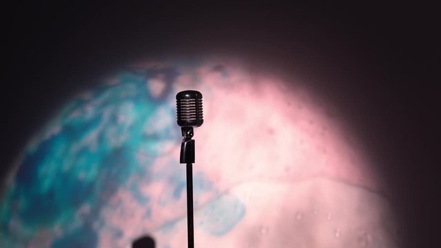 Young Caucasian man in a white unbuttoned shirt goes on stage, adjusts height of classic retro glare microphone. Male picks up chrome mic on scene in the dark club and says 1, 2, 3. Spotlights shine.