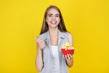 Young beautiful girl with potato chips in bowl on yellow background