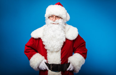 Ho-ho-ho! Real Santa is posing on a blue background with both of his hands on a belt, smiling and looking at the camera.