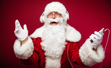 Fototapeta premium Carol of the bells. Authentic Santa Claus is posing with a joyful face expression, holding a smartphone in his left hand and listening to music on the headphones.