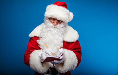 Taking notes. Authentic Santa Claus is posing with a notebook in his left hand, where he writes with the pen, which he holds in his right hand.