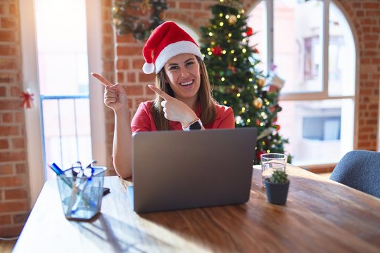 Beautiful woman sitting at the table working with laptop wearing santa claus hat at christmas smiling and looking at the camera pointing with two hands and fingers to the side.