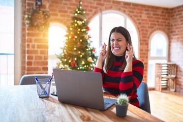 Beautiful woman sitting at the table working with laptop at home around christmas tree gesturing finger crossed smiling with hope and eyes closed. Luck and superstitious concept.