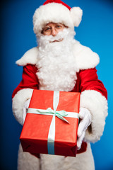 Here you are. Close up photo of a gift, wrapped in a red wrapping paper with a turquoise ribbon on it, which is being held by Santa Claus and showed to the camera by him.