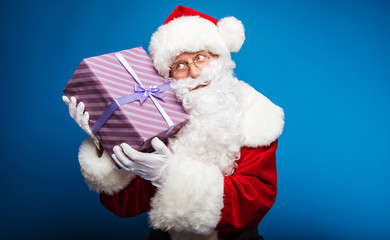 What’s inside? Authentic Santa Claus is holding a striped purple present box near his right ear, smiling and looking to the right.
