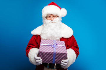 Fototapeta na wymiar Mystery in a box. Close up photo of authentic Santa Claus, who is holding a violet striped present box with a purple ribbon right in front of him, smiling and looking gently at the camera.