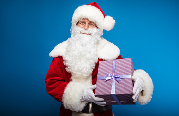 Christmas Eve. Carrying someone&rsquo;s dream. Real Santa Claus is posing with a striped purple gift box, which he is holding in his hands on the left side of him, looking at the camera and smiling kindly.