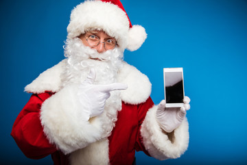 A new device. Santa Claus is holding a smartphone in his hand, showing it to the camera and pointing at it with his right index finger, looking to the camera with his head slightly turned right.