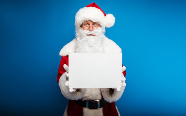 A message. Authentic Santa Claus is posing on a blue background, looking forward, holding a blank postcard and showing it at the camera.