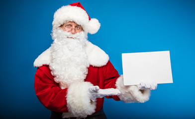 Look! Real Santa is pointing with his right hand at the blank postcard with copyspace on it, which he holds in his left hand, looking at the camera calmly.
