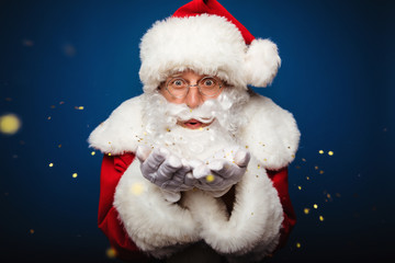 True magic. Close up photo of authentic Santa Claus, blowing a golden confetti off his palms and looking at the camera.