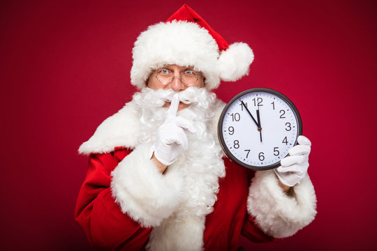 Be quiet! Real Santa Claus is showing a shush sign with his right hand and holding a big round clock which indicate five minutes to twelve in his left hand.