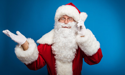 This can’t be! Authentic Santa Claus is posing while talking on the cellphone, which he holds in his left hand, spreading his right hand in surprise.