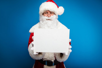 Read this. Close up photo of authentic Santa Claus in round-shaped glasses holding a blank postcard with a copyspace and showing it at the camera.
