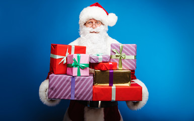Everyone will be pleased. Authentic Santa Claus is holding a pile of multicolored presents in his hands, looking at the camera with a kind smile.