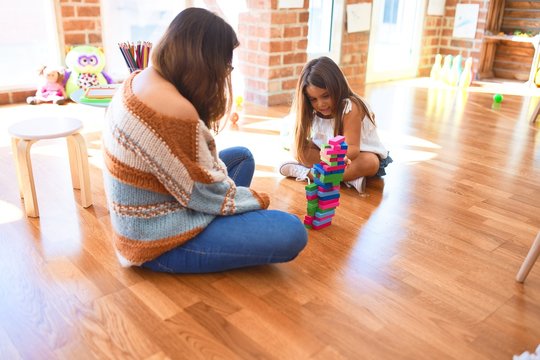 Beautiful teacher and toddler playing with wooden building blocks around lots of toys at kindergarten
