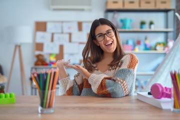 Young beautiful teacher woman wearing sweater and glasses sitting on desk at kindergarten Inviting...