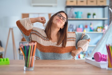 Young beautiful teacher woman wearing sweater and glasses sitting on desk at kindergarten stretching back, tired and relaxed, sleepy and yawning for early morning - Powered by Adobe