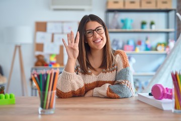 Young beautiful teacher woman wearing sweater and glasses sitting on desk at kindergarten showing...