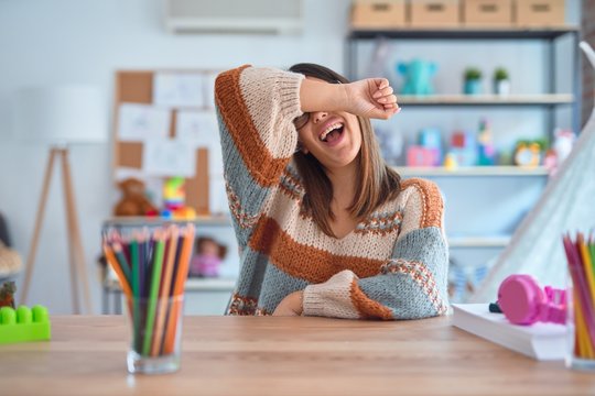 Young beautiful teacher woman wearing sweater and glasses sitting on desk at kindergarten covering eyes with arm smiling cheerful and funny. Blind concept.
