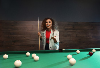 Young African-American woman with cue near billiard table indoors