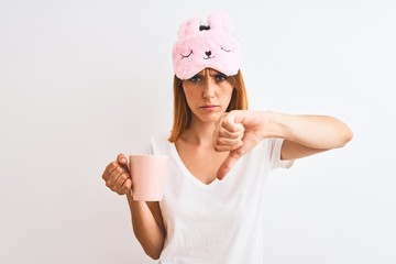 Beautiful redhead woman wearing sleeping mask drinking a cup of coffee over isolated background with angry face, negative sign showing dislike with thumbs down, rejection concept