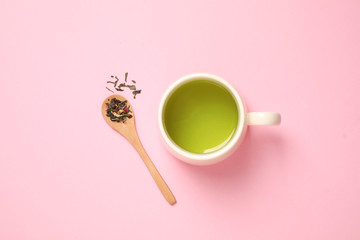 Spoon with green tea leaves and cup of hot beverage on pink background, flat lay