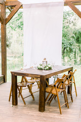 Wedding. Banquet. The wooden chairs and table for guests, decorated with flowers and greenery, served with cutlery and wineglasses. The table stands on a wooden floor in reastaurant outdoors in forest